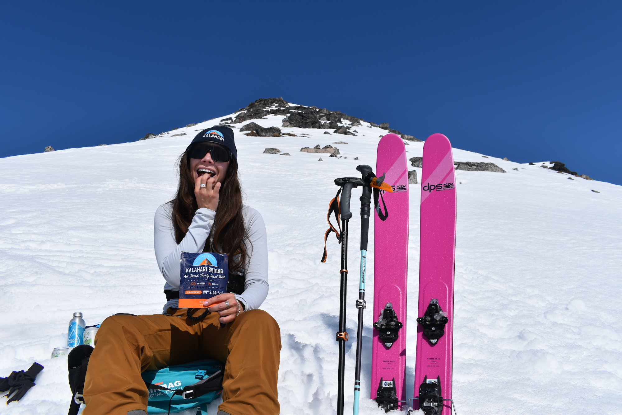 woman sits on snow covered mountain eating biltong with bright pink skis standing next to her
