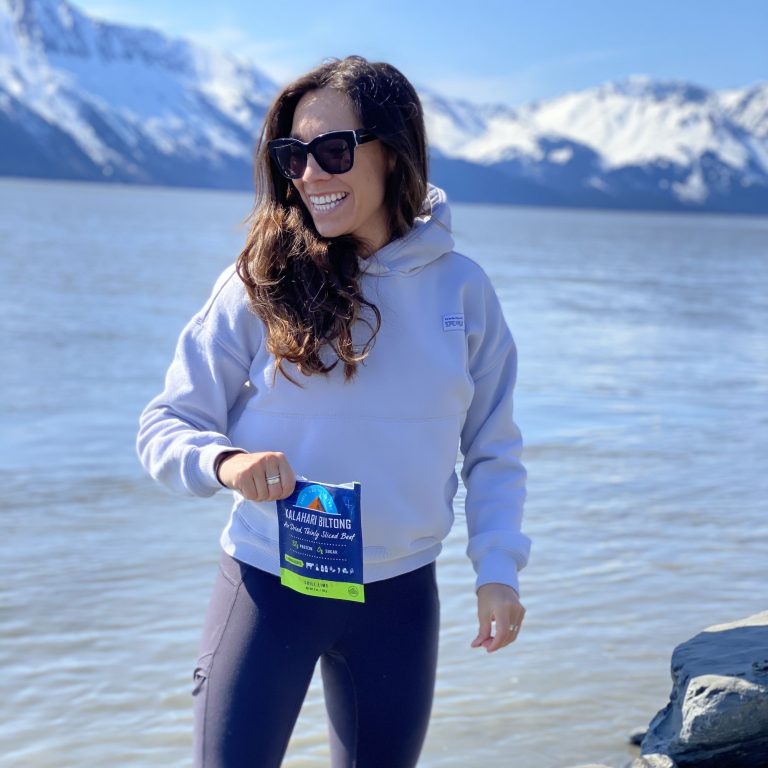 woman with long brown hair in grey hoodie and leggings standing smiling with biltong bag in front of lake in front of snow capped mountains
