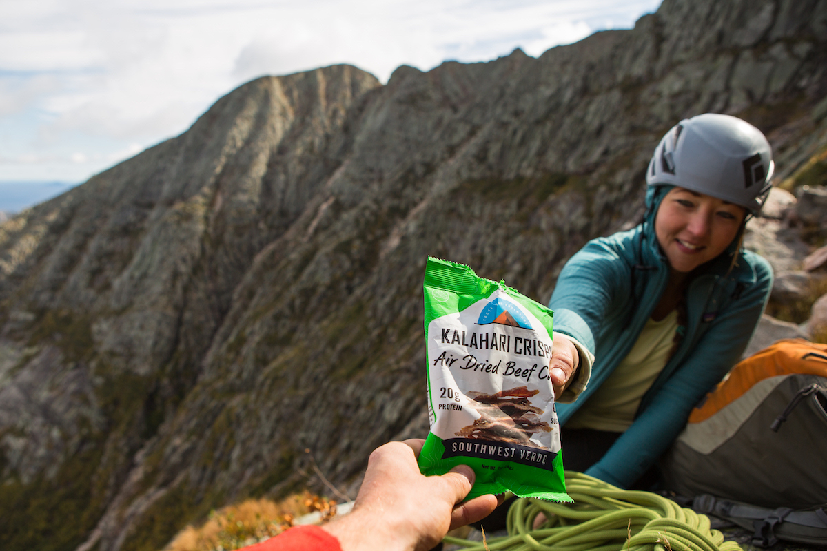 woman in climbing gear and helmet reaching for Southwest Verde keto crisps with mountain in the background