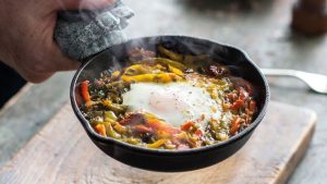 Hand holding cast iron pan cooking egg, veggies, Biltong on top