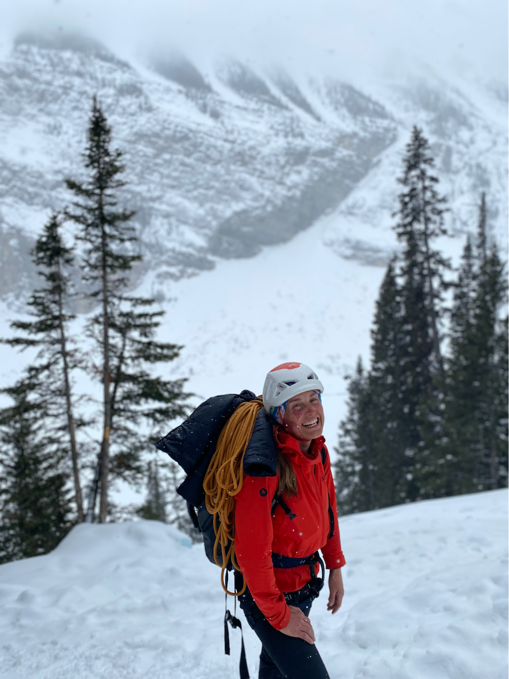 smiling woman in red jacket, climbing helmet, with climbing pack in snowy mountains